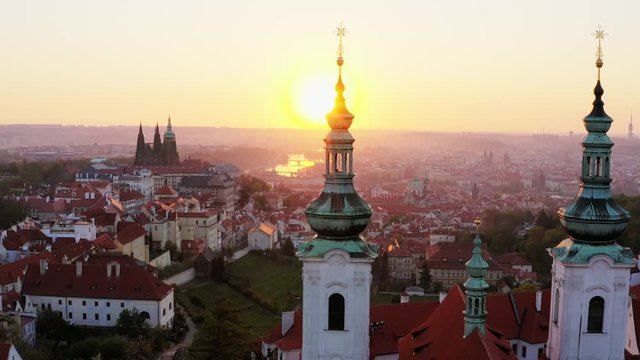 Aerial view over Strahov monastery at sunrise. Lateral shot, drone circling near belfries of the cathedral decorated crucifix on spire in sun shining, showing picturesque panorama above Prague