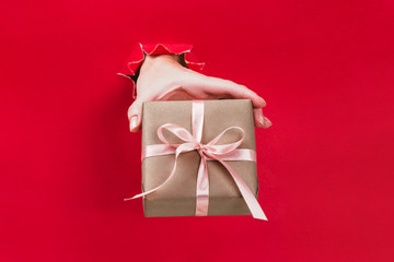 Female hand holding gift box wrapped in craft paper with pink ribbon through a hole in red...