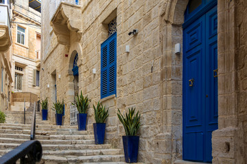Narrow charming street in Birgu, Malta, with limestone medieval buildings with navy blue doors and window shutters and plants in blue pots along the walls.