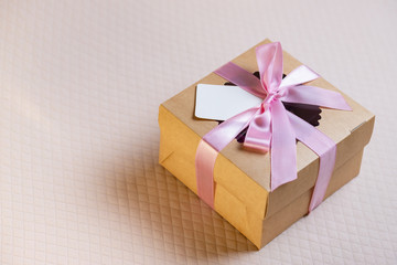 A square gift box with a pink ribbon on a light pink background. Cupcakes inside. Space for text, empty note tied over.