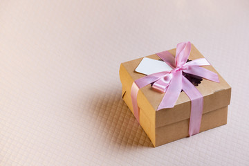 A square gift box with a pink ribbon on a light pink background. Cupcakes inside. Space for text, empty note tied over.