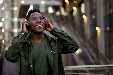smiling african-american man in stylish jacket in wireless headphones listening music on the street of the evening city. space for text