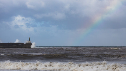 Olas rompiendo contra un faro durante un temporal con el arco iris en el cielo