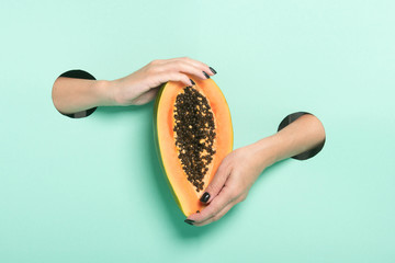 Female hands hold fresh papaya through a hole on neon mint background.
