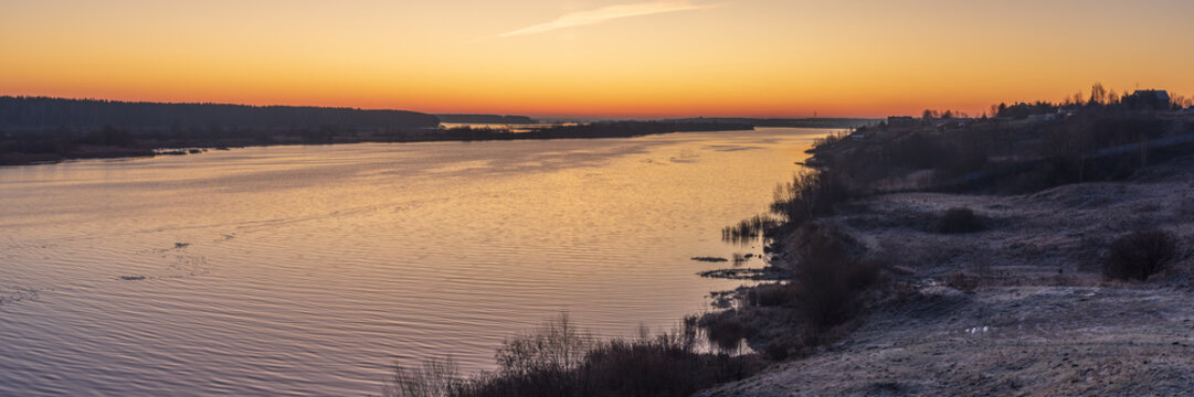 Panoramic view of the golden sunrise over a large river covered with the first ice. November, earlier morning