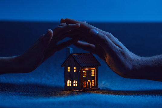 home protection. male and female hand on a small house in which the windows glow with warm light