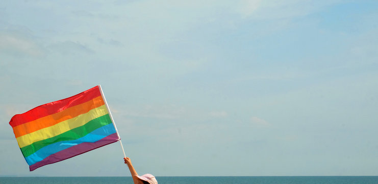A hand holding an LGBTQ rainbow flag flying in the sky, the flag is in the bottom right of the image and the rest is sky, ideal for copy space