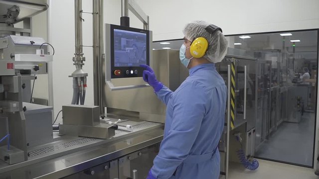 Man employee work with computer control production process in pharmaceutical factory. Portrait of american lab worker in protective clothes is watching and touching pc screen standing in room of plant
