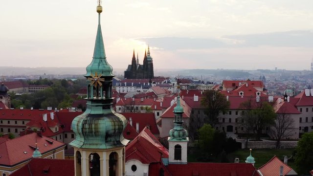 View cropped cathedral tower over red tiled roof drone moving to tower decorated symbol of christianity. Aerial panorama above Loreta cloister and old history centre of Prague at sunrise 
