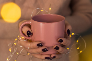 A girl in a delicate pink sweater with dark manicure holds a mug of warm drink in her hands with a yellow garland. The atmosphere of Christmas, New Year, warmth and lights of a garland