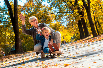 Smiling granddaughter enjoying day with grandmother in the autumn park while riding skateboard