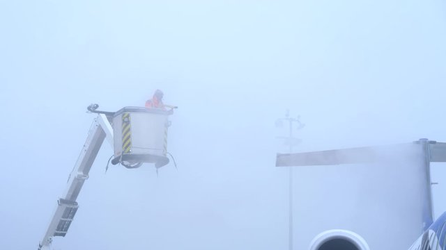An aircrafts tail is being deiced by a man spraying fluid from an aerial platform. Removal of frost prior the flight. Misty day. Handheld.