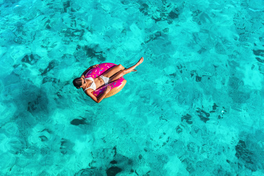 Beach vacation woman relaxing in pool float donut inflatable ring floating on turquoise ocean water background in Caribbean travel summer. Girl in white bikini top drone view.