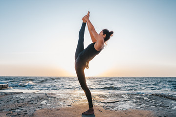 Fototapeta na wymiar Young woman with curly hair in black costume practicing yoga at sunrise light, minimalist scene. Doing asana. Healthy lifestyle concept.