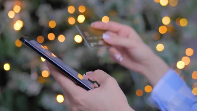 Woman using smartphone, holding credit card, buying goods or ordering online. Garland light bokeh background. Christmas, e-commerce, sale, consumerism, electronic payment, online shopping concept
