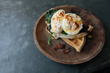 Close up Breakfast with Poached Egg on waffle with spinach and bacon on wooden texture plate on grey stone table. Top view