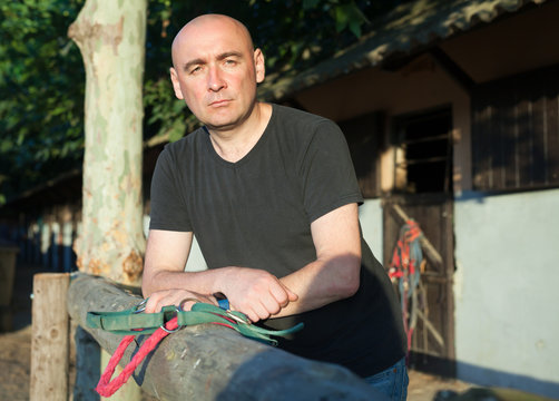 Mature smiling man holding a surcingle and standing at farm