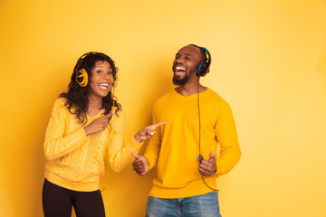 Young emotional african-american man and woman in bright casual clothes on yellow background. Beautiful couple. Concept of human emotions, facial expession, relations. She's pointing on singing man.