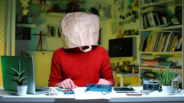 Self-isolation at home, during quarantine of coronavirus infection COVID-19. Crazy sick man sitting at a table in the office. On his head is a paper bag.