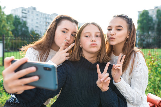 Three teenage girls. They take pictures themselves on phone, happy smiles play and fool around. Online application on Internet, social networks. In summer city, warm autumn sweaters.