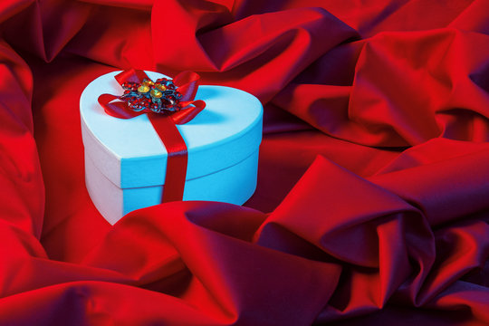 valentines greeting card in blue light. white cardboard box in shape of heart wrapped in ribbon lay on a red cloth which repeats the form of present package. love and romance gift concept