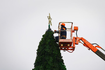 Municipal workers on height in crane bucket install star on top of Christmas tree. Worker hang star and decorate pine. Preparation for New year celebration. Construction of the city Christmas tree