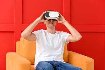 Young man with virtual reality glasses sitting in armchair on color background