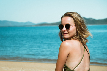 Young attractive girl with sunglasses sitting on the beach, getting tanned, looking to the camera