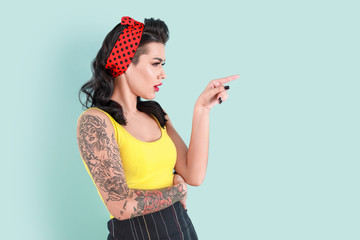 Portrait of beautiful tattooed pin-up woman pointing at something on color background