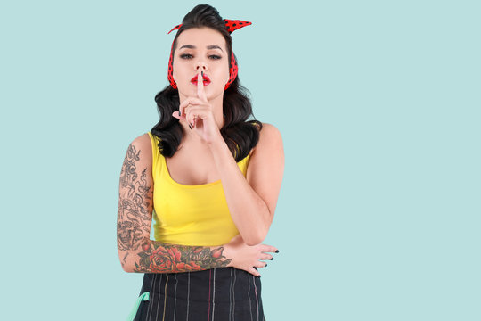 Portrait of beautiful tattooed pin-up woman showing silence gesture on color background