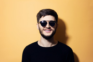 Fototapeta premium Portrait of young smiling hipster, dressed in black, wearing sunglasses, isolated on yellow background.