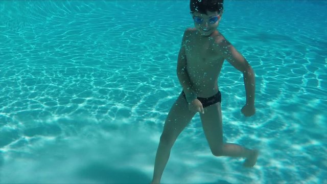 Funny cheerful boy in swimming goggles dives under the water in the pool and dances happy dance
