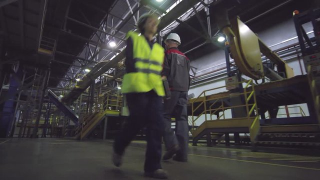 Full rear tracking shot of male engineer in overalls and hardhat, with roll of technical papers, walking along mineral wool production line at manufacturing plant, and female colleague passing by