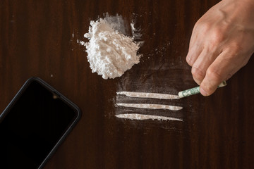 Drug Abuse. Hand of man holds rolled banknote for snorting line of cocaine powder and mobile phone on the table - obrazy, fototapety, plakaty
