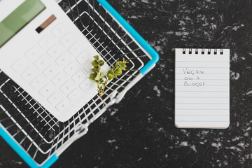 vegan on a budget text on notepad with shopping basket calculator and branch of leaves