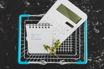 vegan on a budget text on notepad with shopping basket calculator and branch of leaves