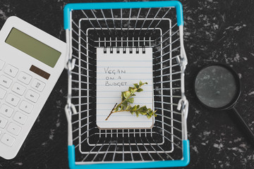 vegan on a budget text on notepad with shopping basket calculator and branch of leaves