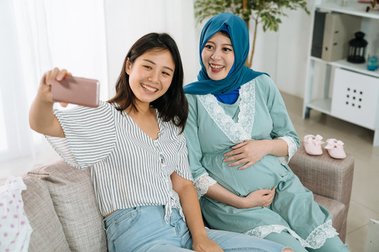 Beautiful pregnant woman and friend making selfie using smart phone and smiling while celebrating baby shower in cozy apartment. muslim young future motherhood in blue hijab and taking self portrait