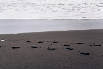 Footprints on black sand beach shore by ocean with sea foam