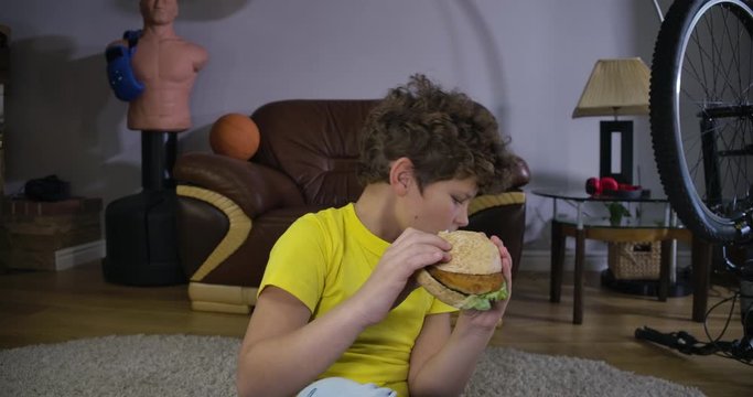 Absorbed Caucasian teenage boy watching TV, chewing hamburger, starting coughing. Portrait of cute teenage kid with junk food at home. Leisure activity, relaxation, generation Z. Cinema 4k ProRes HQ.