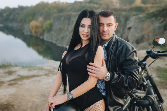 Stylish man biker hugs a beautiful girl in a black dress and stockings sitting on a modern black motorcycle. Portrait of a couple in love on nature in cloudy weather. Photography, concept, love story.