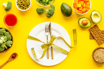Diet concept. Fork and knife tied with measure tape on plate near vegetables on yellow background top view