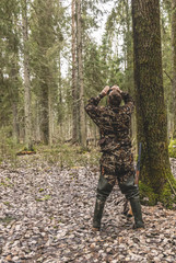 a man in camouflage in the forest, autumn, stands by a tree and looks through binoculars