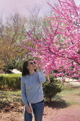 tender girl in a blue sweatshirt and blue jeans, round glasses, enjoys the bloom of a rosewood on a bright sunny day outdoors