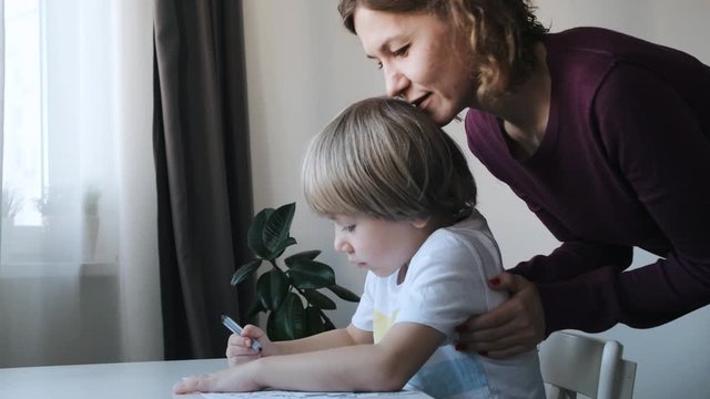 little boy, kid, sitting at the table at home and drawing, mother kissing and hugging him