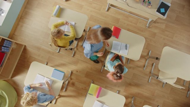 Bright Elementary School Classroom: Children Sitting At Their School  Desk Working, Doing Assignment, Kind Teacher Walks Between Rows Helping Kids With Homework Tasks. Spinning Top View Camera Shot.