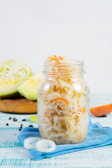 A jar of fermented cabbage stands on a wooden table with fresh vegetables around. Vertical orientation.
