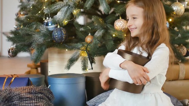 Smiling cute blond girl is hugging a gold gift box she recieved sitting near christmas tree, happy childhood and xmas celebration concepts