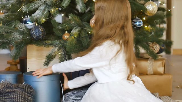 Cute blond girl is opening dark blue gift box she recieved sitting near christmas tree, happy childhood and xmas celebration concepts