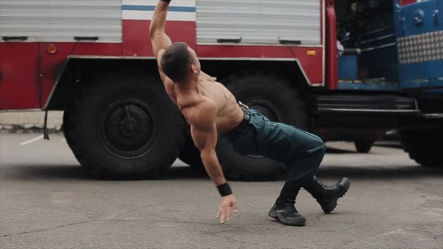 Slow motion. Attractive shirtless young athlete exercising push-ups with jumping. Big fire engine in the background. Side view